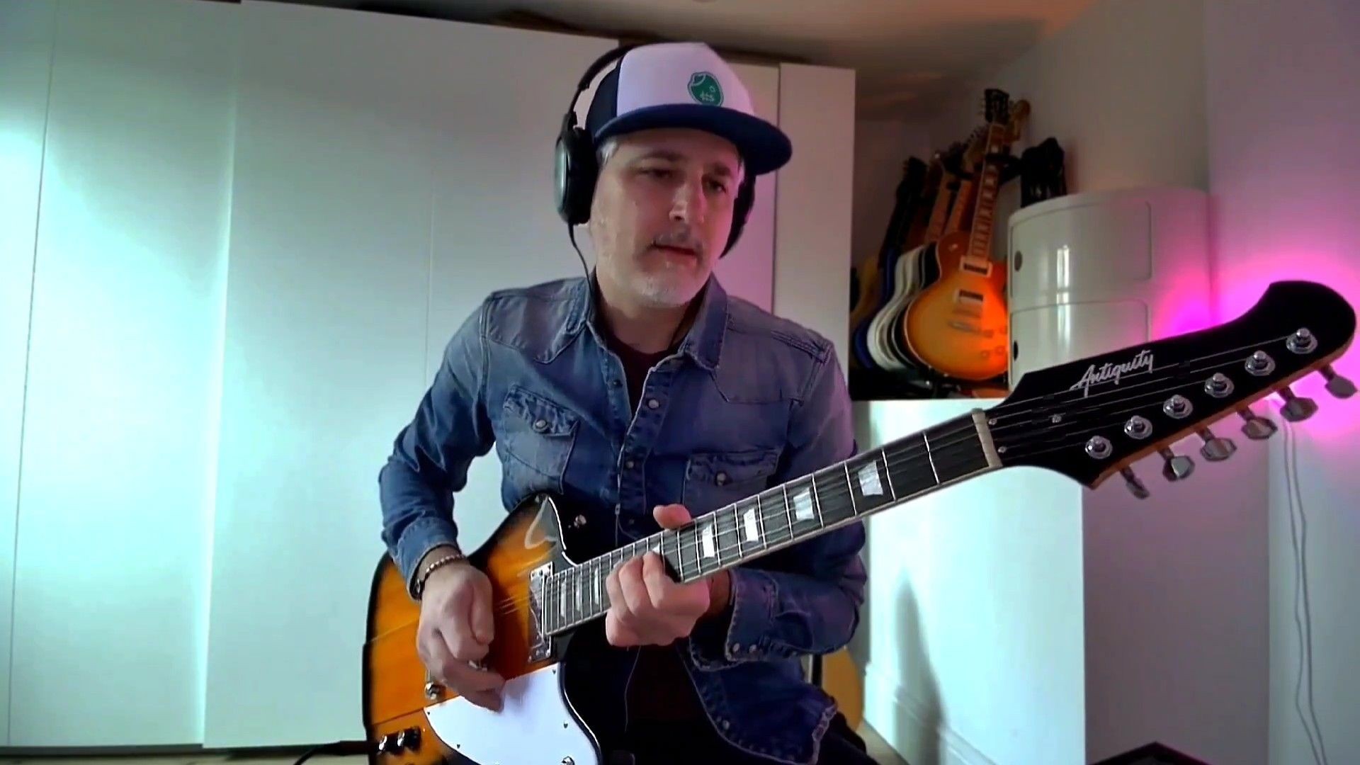 Man in headphones and denim jacket playing electric guitar in a room with guitars hanging on the wall.