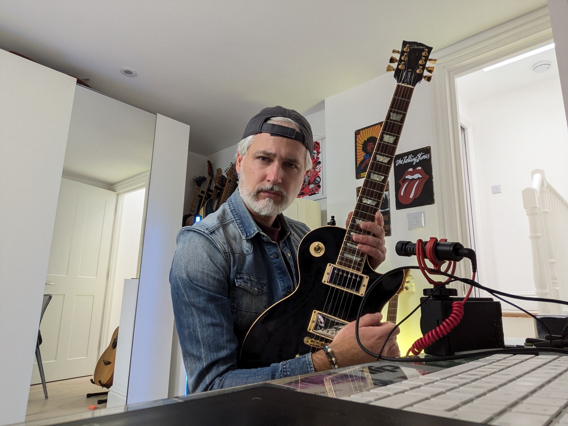 Man in denim shirt holding a black electric guitar in a music-themed room with posters and equipment.